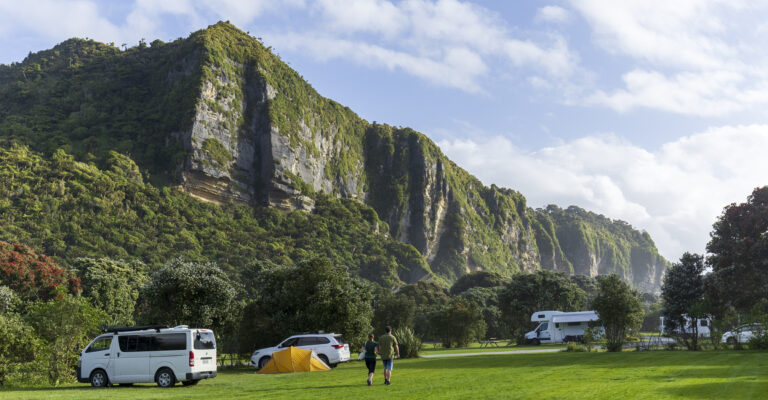 a landscape image of Punakaiki beach camp showing the blue sky, and cliff side with green grass and guests walking through camp with a car, motorhome and tent set up